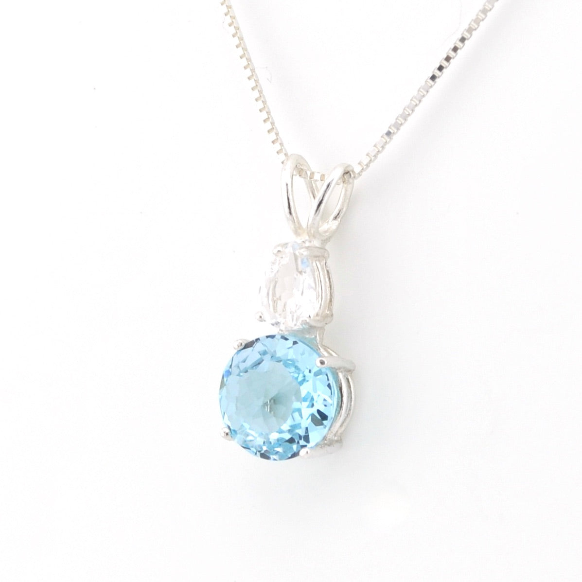 A silver pendant necklace featuring a round light blue topaz gemstone and a small pear-cut clear crystal on a delicate silver chain, set against a plain white background.