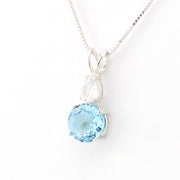 A silver pendant necklace featuring a round light blue topaz gemstone and a small pear-cut clear crystal on a delicate silver chain, set against a plain white background.