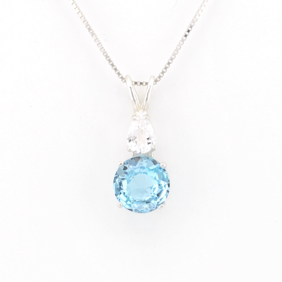 A silver pendant necklace featuring a round light blue topaz gemstone and a pear-cut clear stone, displayed on a clean white background.