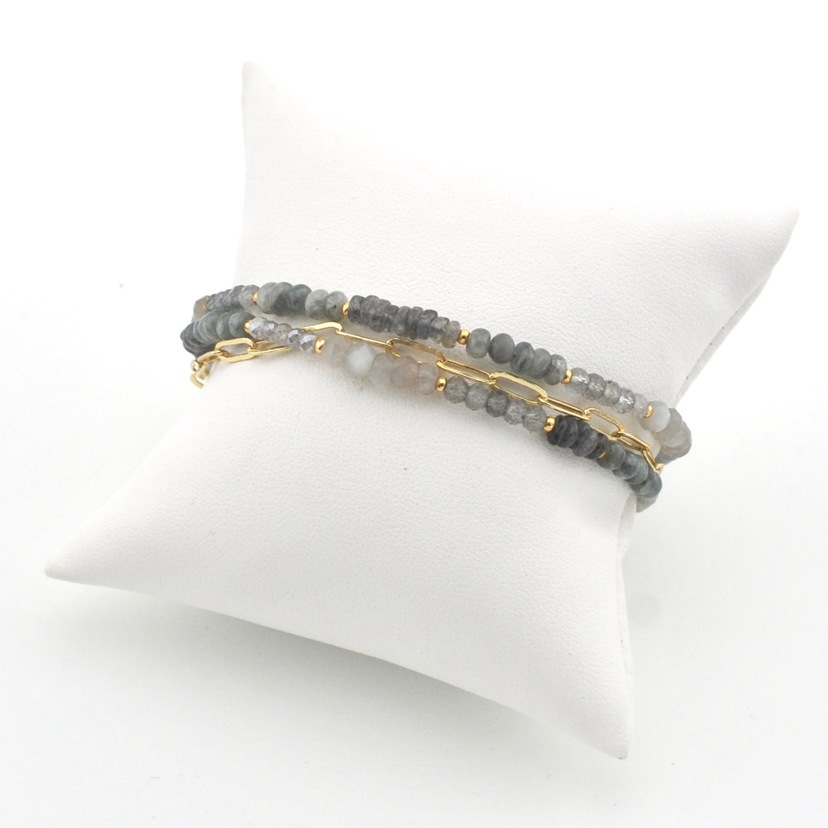 A pair of beaded bracelets with gray stone beads and gold chain accents, displayed on a white pillow against a white background.