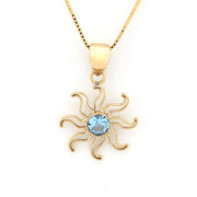 A gold pendant necklace featuring a stylized sun design with a round light blue gemstone in the center, suspended from a gold chain on a white background.