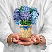 Person holding a small woven basket with blue flowers and a blue ribbon against a white background