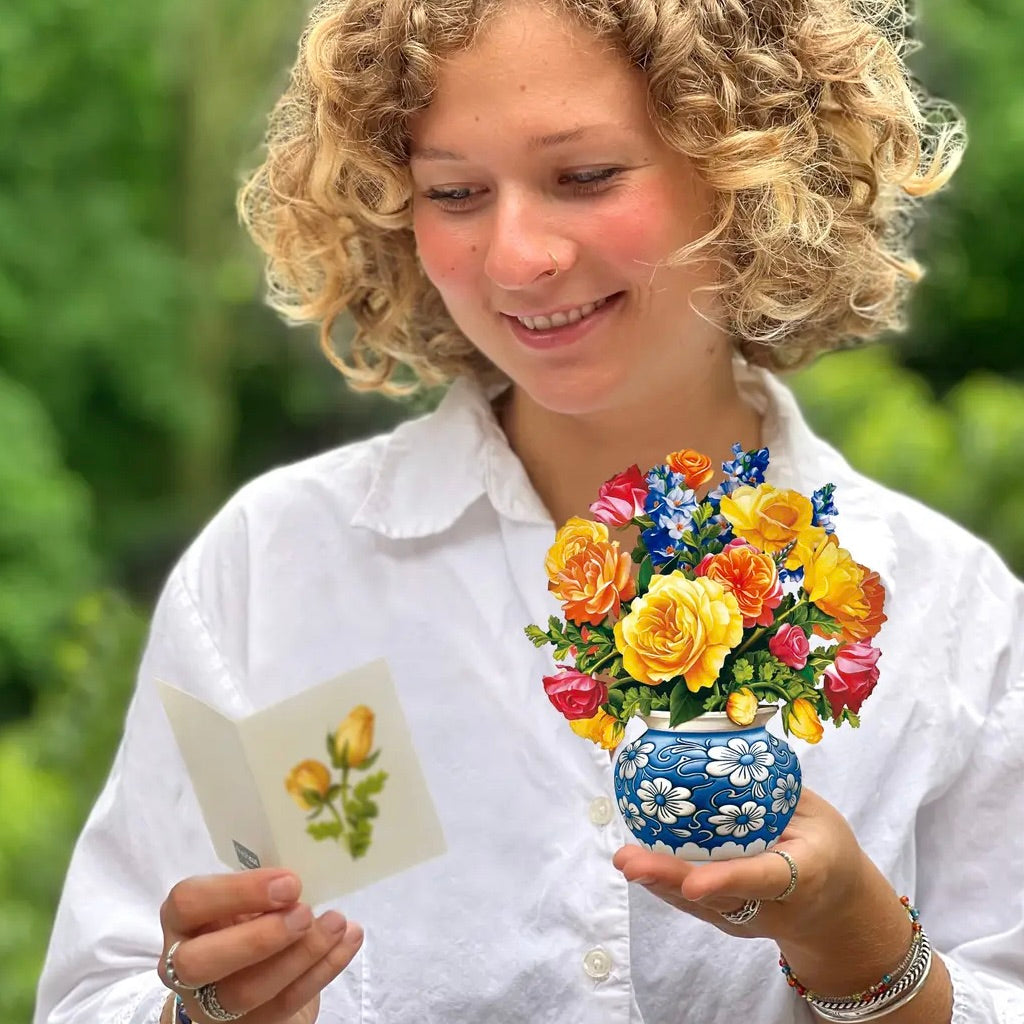 Woman holding a colorful flower arrangement and a card with flowers outdoors.