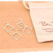 Set of silver square earrings on a beige surface with a branded pouch.
