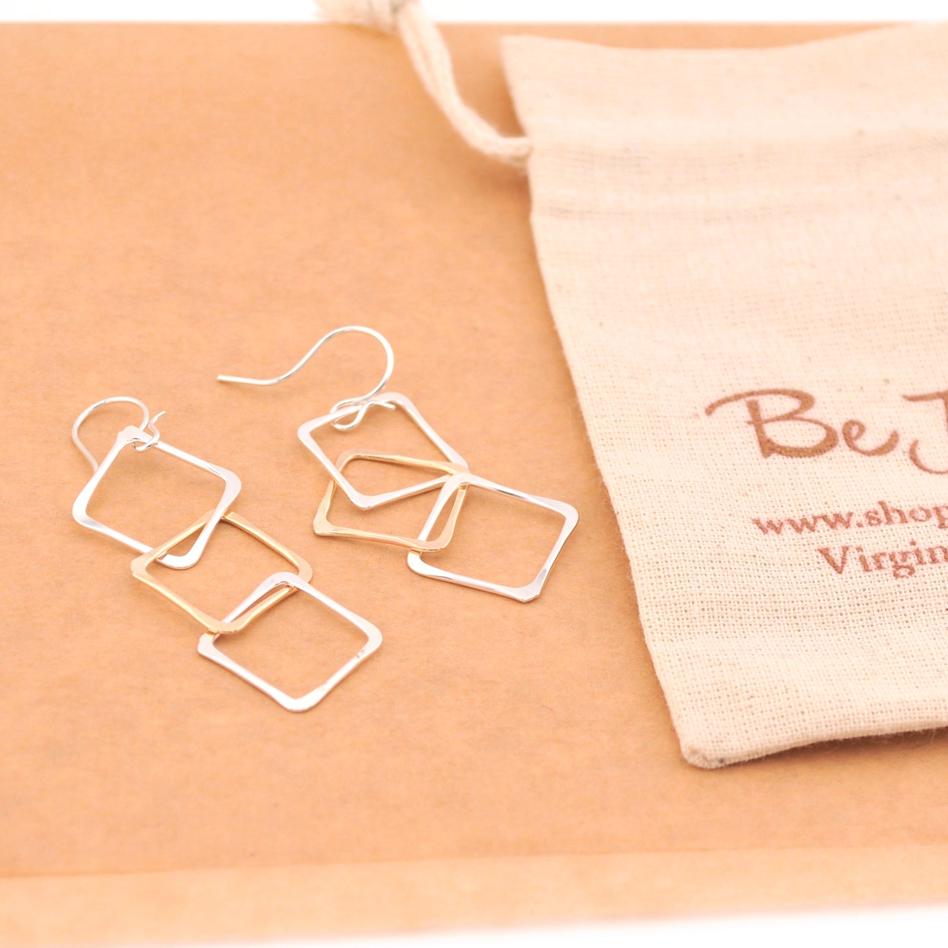 Set of silver square earrings on a beige surface with a branded pouch.