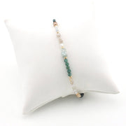 A delicate beaded stretch bracelet displayed on a white cushion. The bracelet features a central strand of green beads, light blue beads, clear beads, and small gold spacer beads.