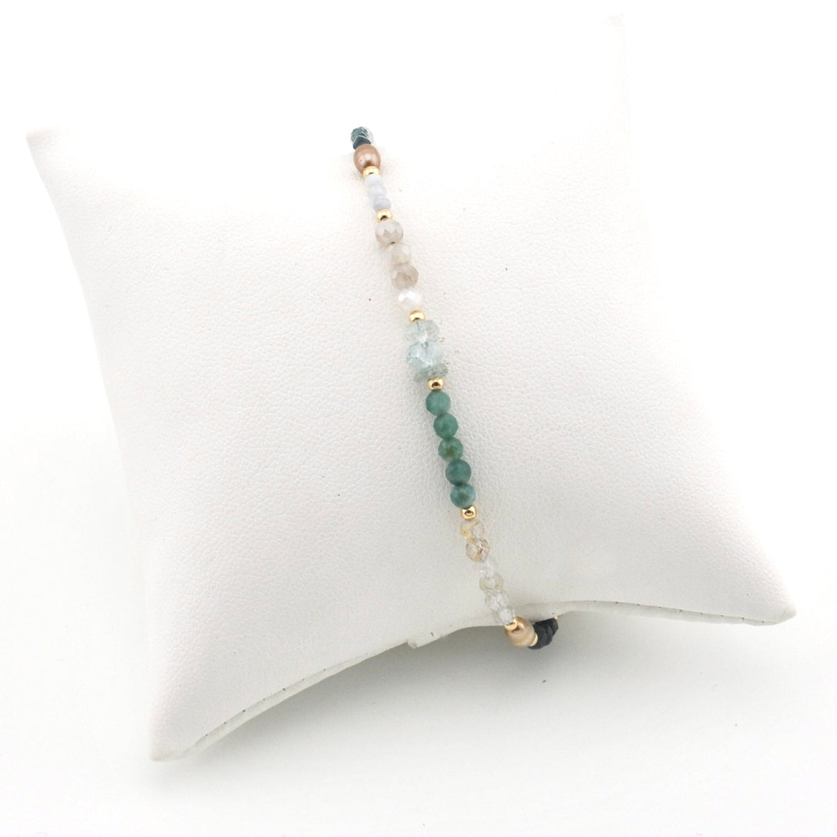 A delicate beaded stretch bracelet displayed on a white cushion. The bracelet features a central strand of green beads, light blue beads, clear beads, and small gold spacer beads.