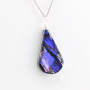 A teardrop-shaped pendant necklace featuring a vibrant blue and purple dichroic glass with pink and green shimmer, set in a silver bezel on a silver chain against a plain white background.