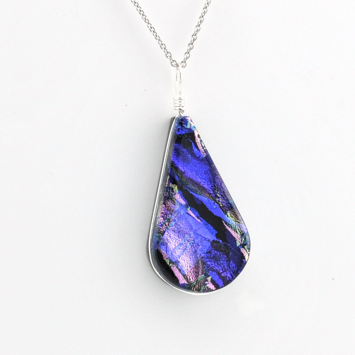 A teardrop-shaped pendant necklace featuring a vibrant blue and purple dichroic glass with pink and green shimmer, set in a silver bezel on a silver chain against a plain white background.