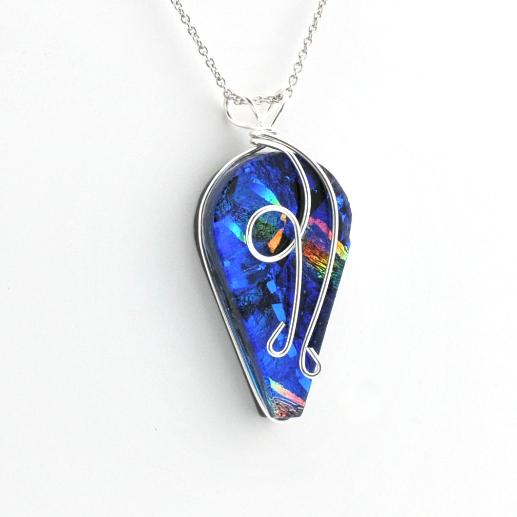 A silver wire-wrapped pendant necklace featuring a vibrant blue iridescent dichroic glass with rainbow reflections, suspended from a delicate silver chain against a plain white background.