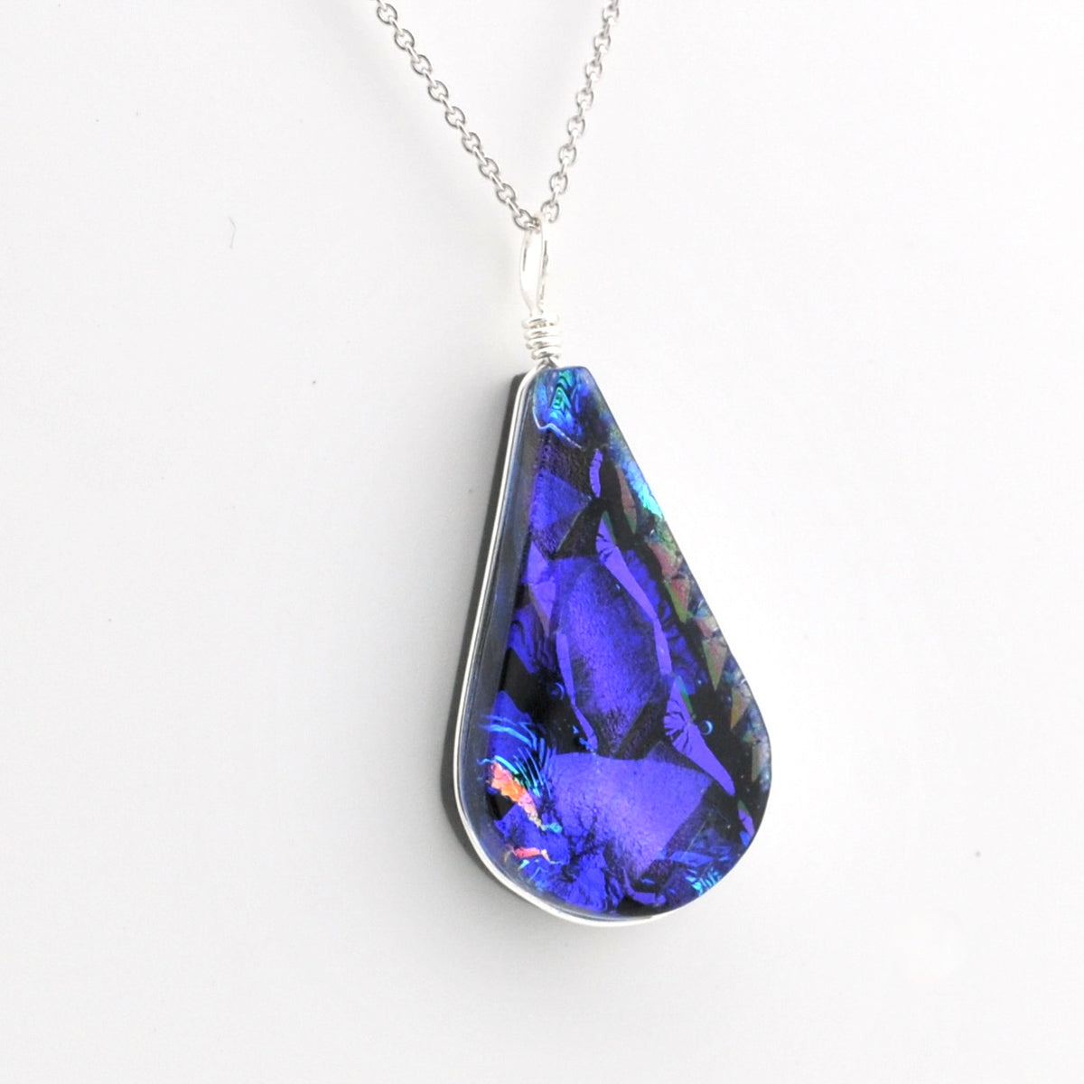 A teardrop-shaped pendant necklace featuring a vibrant blue and purple iridescent dichroic glass set in a silver bezel, suspended from a delicate silver chain against a plain white background.