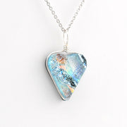 A silver necklace featuring a heart shaped pendant filled with iridescent blue, teal, and orange glass, shown on a clean white background.