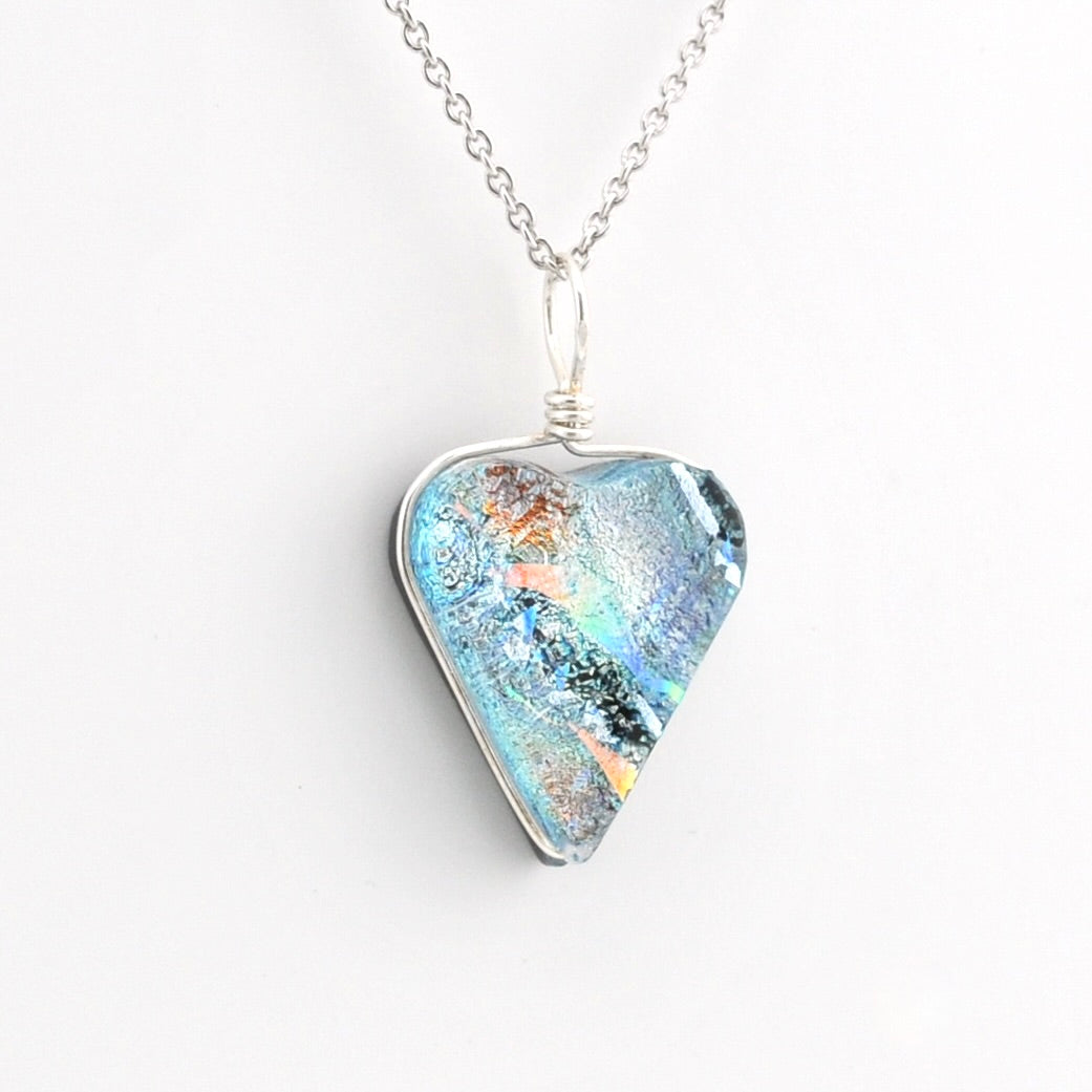 A silver necklace featuring a heart shaped pendant filled with iridescent blue, teal, and orange glass, shown on a clean white background.