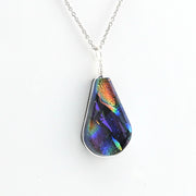 A teardrop-shaped pendant necklace featuring an iridescent, dichroic glass that shifts between purple, blue, green, orange, and gold, suspended from a silver chain on a plain white background.