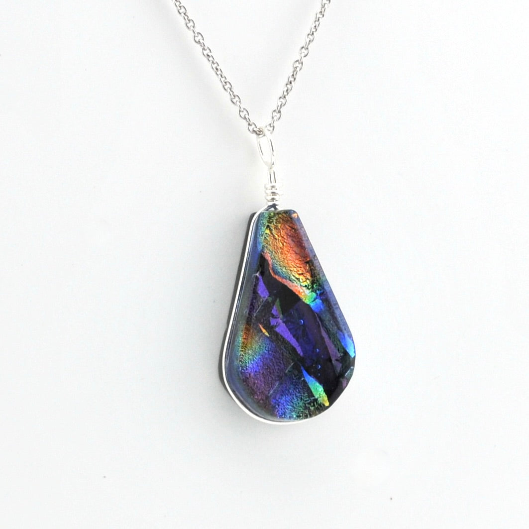 A teardrop-shaped pendant necklace featuring an iridescent, dichroic glass that shifts between purple, blue, green, orange, and gold, suspended from a silver chain on a plain white background.