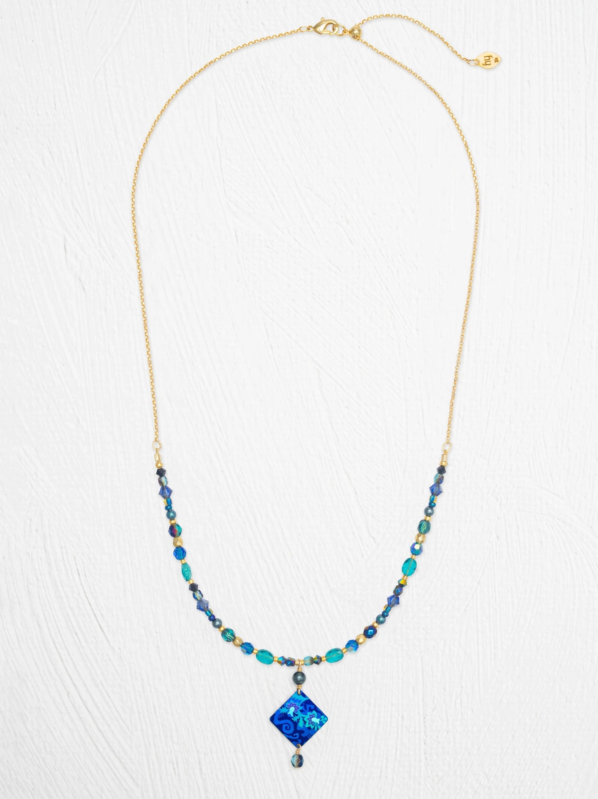 A gold chain necklace featuring a central blue diamond-shaped pendant and beaded strands of blue, teal, and dark gray beads, displayed on a textured white background.