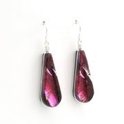 A pair of teardrop-shaped glass dangle earrings with a metallic pink and purple iridescent finish, suspended from silver fishhook wires on a plain white background.