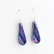A pair of teardrop-shaped dangle earrings featuring iridescent purple and blue dichroic glass set in silver, hanging from silver fishhook wires on a plain white background.