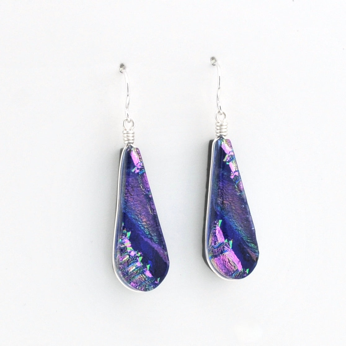 A pair of teardrop-shaped dangle earrings featuring iridescent purple and blue dichroic glass set in silver, hanging from silver fishhook wires on a plain white background.