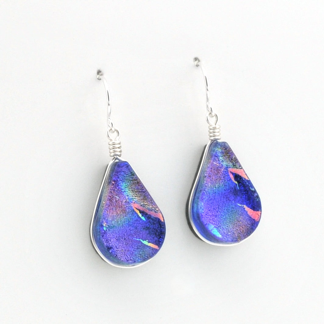 A pair of teardrop-shaped glass drop earrings with a vibrant blue, purple, and pink iridescent pattern, suspended from silver fishhook wires on a plain white background.