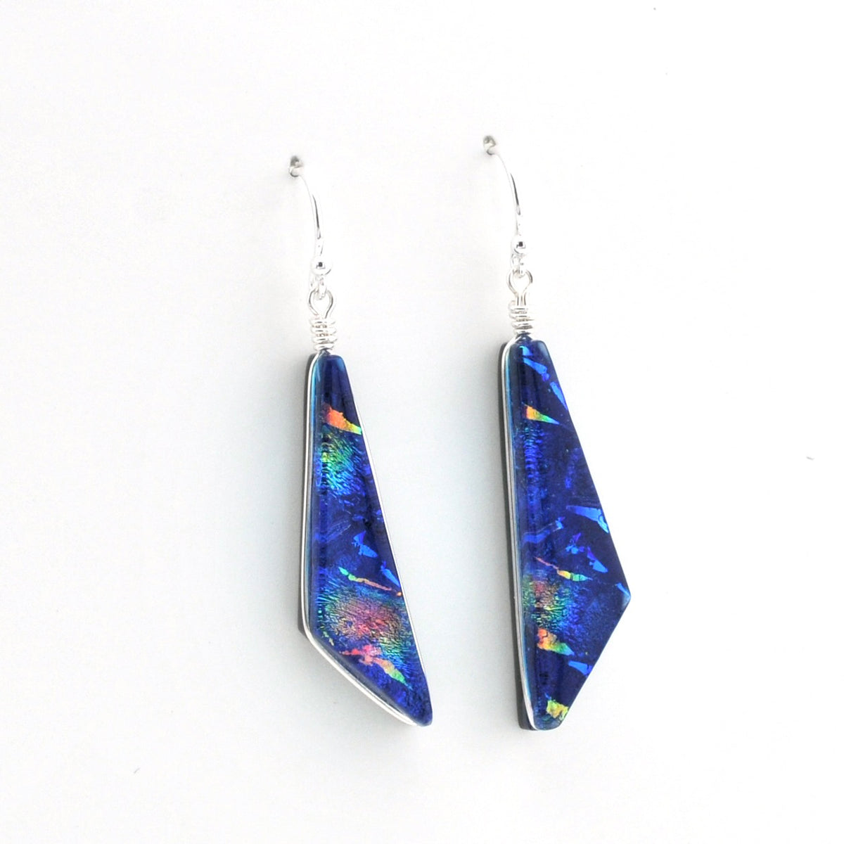 A pair of silver dangle earrings featuring elongated, faceted blue glass with vibrant iridescent flashes of green, orange, and pink, suspended from silver fishhook wires on a plain white background.