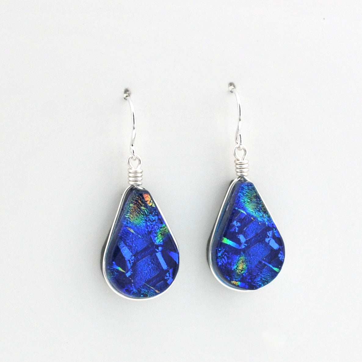 A pair of teardrop-shaped dangle earrings featuring vibrant blue iridescent dichroic glass set in silver bezel settings with silver fishhook wires, shown against a plain white background.