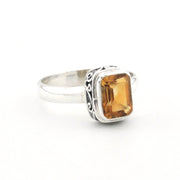 A silver ring featuring a rectangular faceted amber-orange gemstone in a bezel setting with decorative scrollwork on the band, shown on a plain white background.