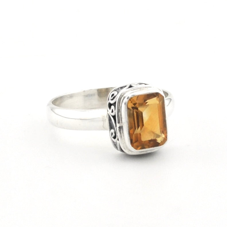 A silver ring featuring a rectangular faceted amber-orange gemstone in a bezel setting with decorative scrollwork on the band, shown on a plain white background.