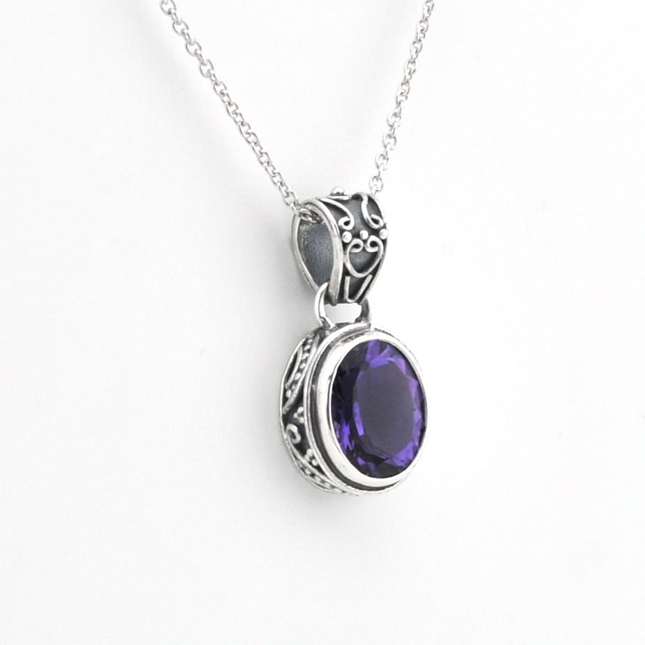 A silver pendant necklace featuring an oval-cut deep purple gemstone in an ornate silver bezel setting, suspended from a delicate silver chain against a plain white background.