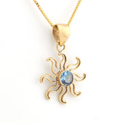 A gold pendant necklace featuring a stylized sun design with a central light blue gemstone, shown on a clean white background.