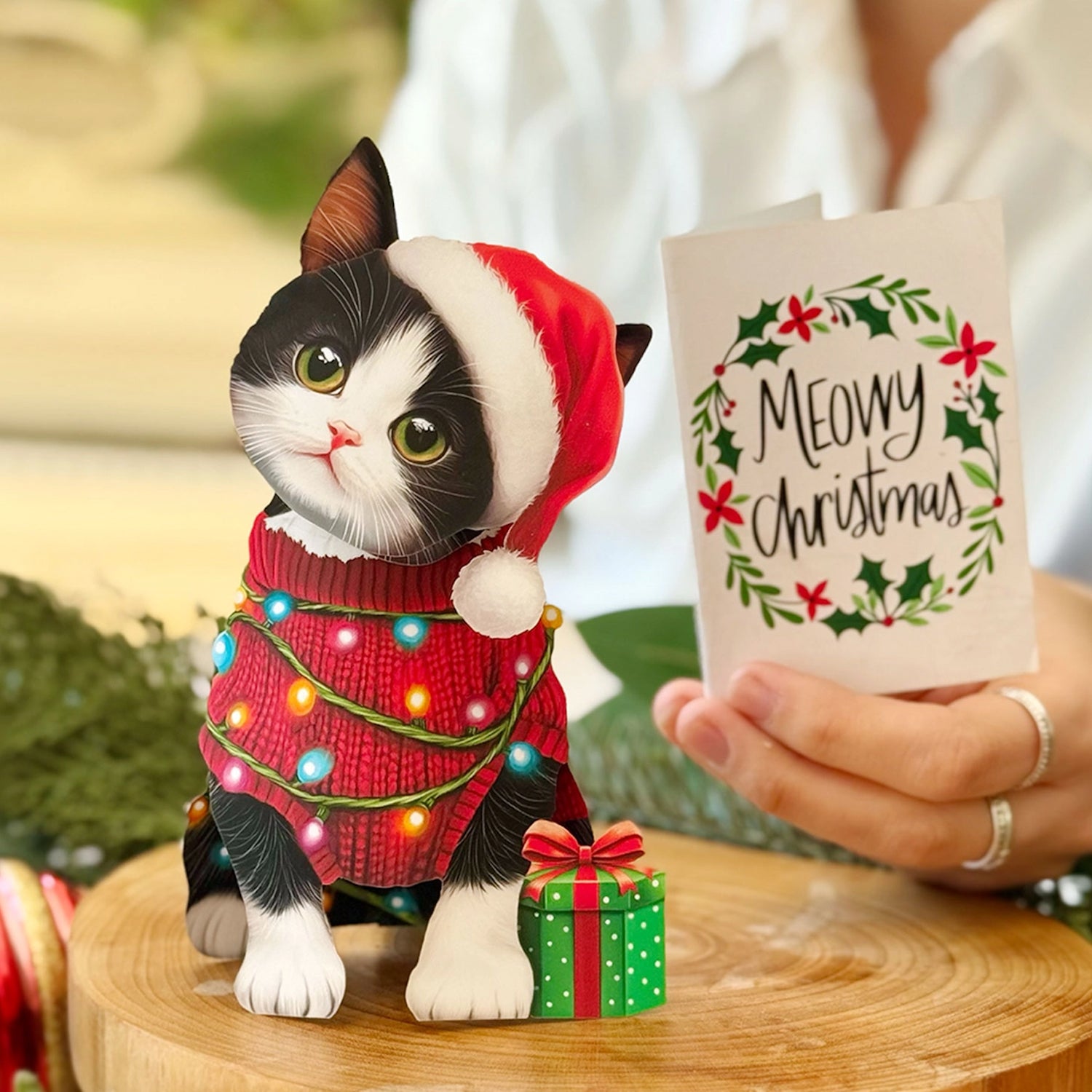 Cat figurine in a Santa hat with Christmas lights and a gift, held by a person with a 'Meowy Christmas' card.