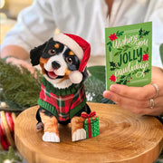Dog figurine in a Santa hat and plaid sweater with a Christmas card on a wooden surface.