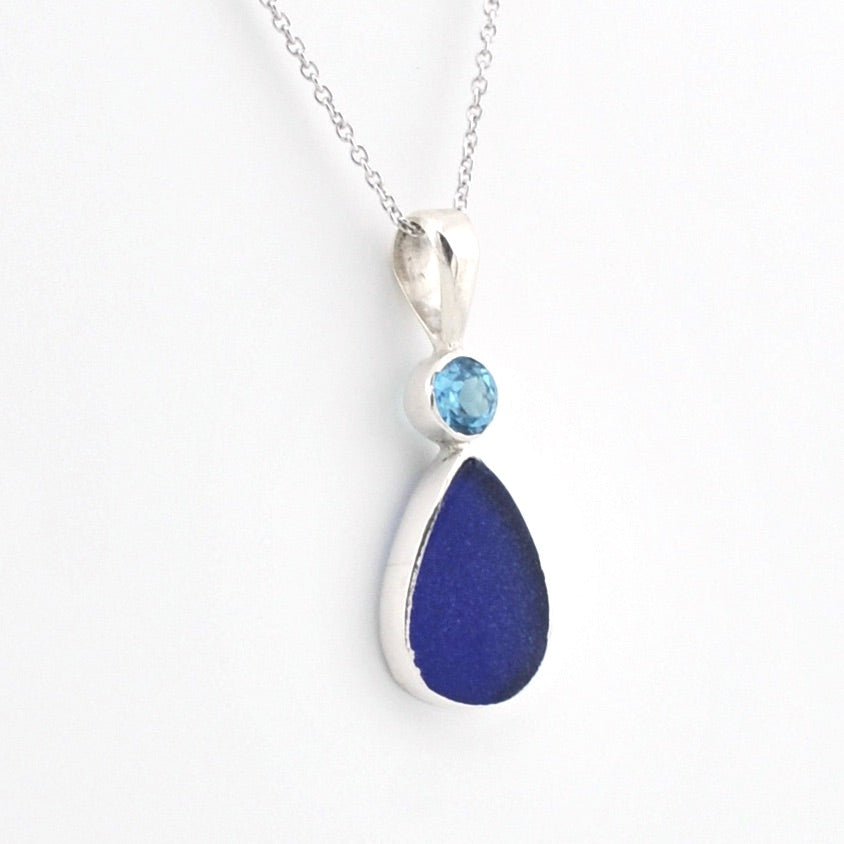 A silver pendant necklace featuring a teardrop-shaped dark blue glass and a small round light blue gemstone, shown on a white background.