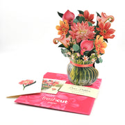 Bouquet of flowers in a glass vase with a pink ribbon, placed on a pink card with 'fresh cut paper' branding.