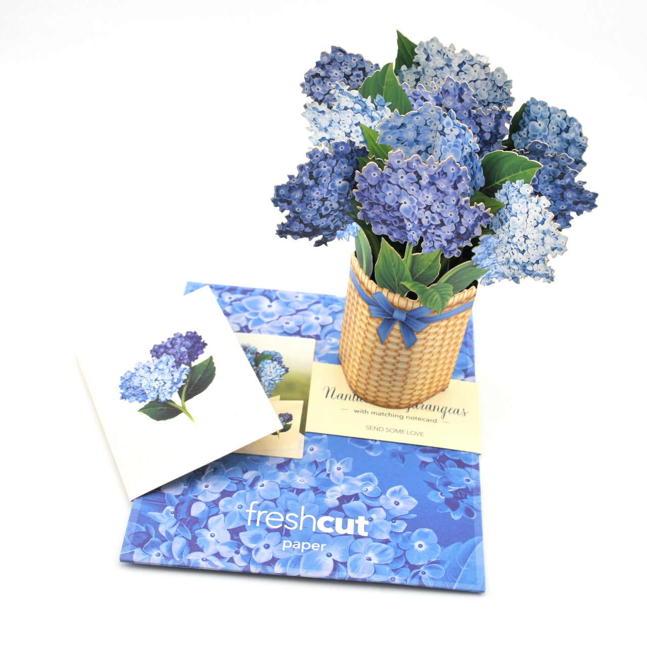 Bouquet of blue flowers in a woven basket with a card on a white background, featuring 'freshcut paper' branding.