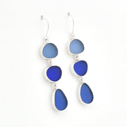 A pair of silver drop earrings featuring three stacked oval and teardrop-shaped sea glass stones in shades of blue, set against a plain white background.