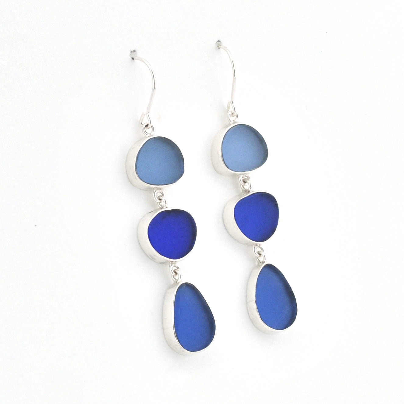 A pair of silver drop earrings featuring three stacked oval and teardrop-shaped sea glass stones in shades of blue, set against a plain white background.