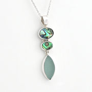 A silver pendant necklace featuring three stacked oval and teardrop-shaped iridescent green abalone shell pieces and a large smooth sea glass drop, shown on a plain white background.