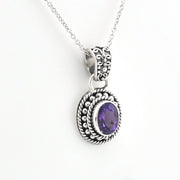 A silver-toned oval pendant necklace featuring a textured, ornate filigree border and a central bezel-set purple amethyst gemstone, suspended from a delicate silver chain against a plain white background.
