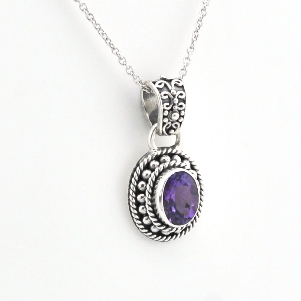 A silver-toned oval pendant necklace featuring a textured, ornate filigree border and a central bezel-set purple amethyst gemstone, suspended from a delicate silver chain against a plain white background.