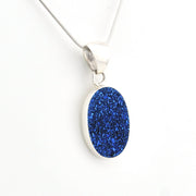 A silver pendant necklace featuring an oval-shaped, glittery royal blue stone, suspended from a delicate silver chain against a plain white background.