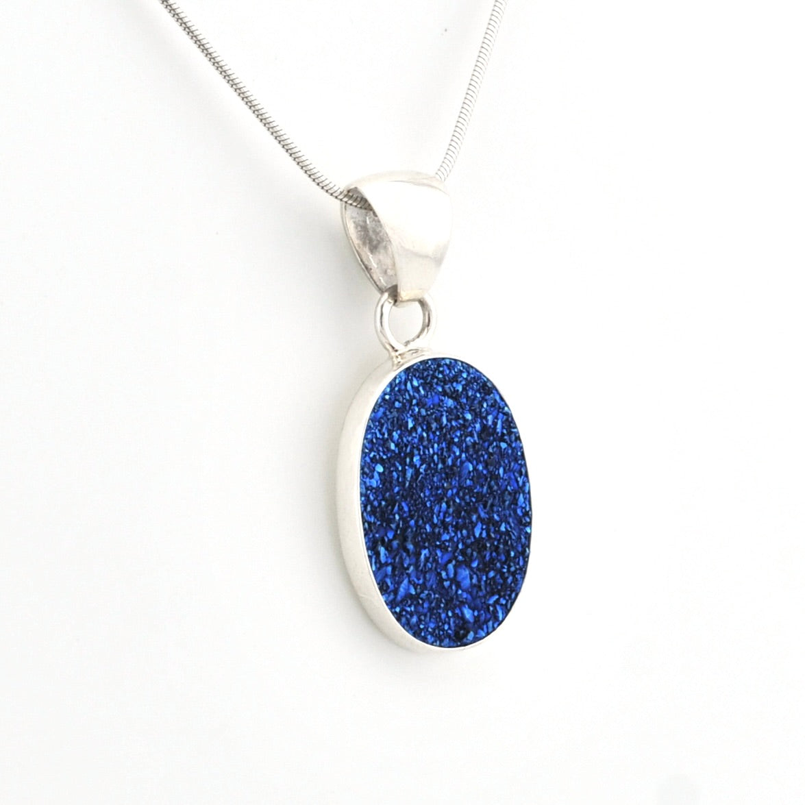 A silver pendant necklace featuring an oval-shaped, glittery royal blue stone, suspended from a delicate silver chain against a plain white background.
