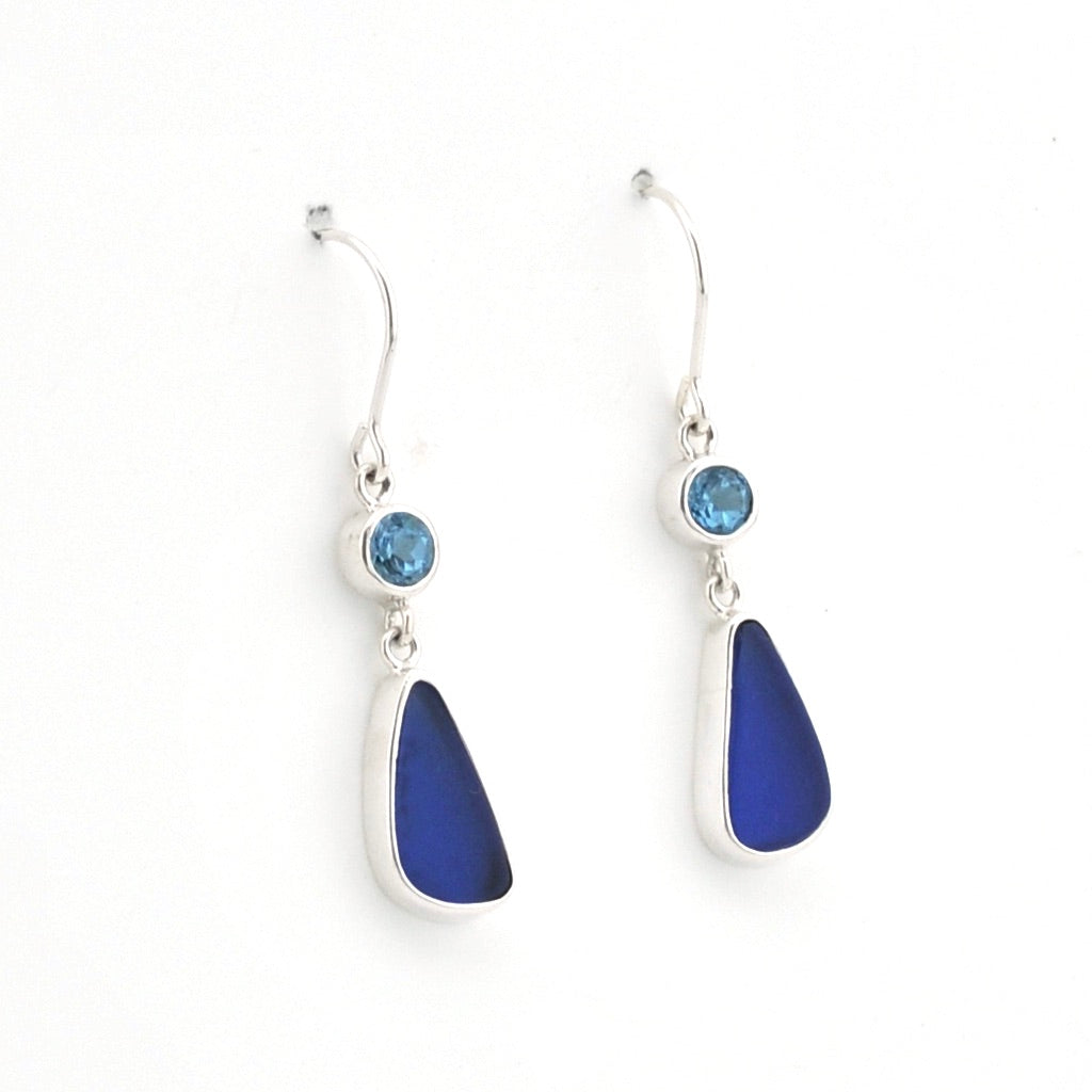 A pair of silver drop earrings featuring a small round light blue gemstone and a larger teardrop-shaped deep blue sea glass, shown on a plain white background.
