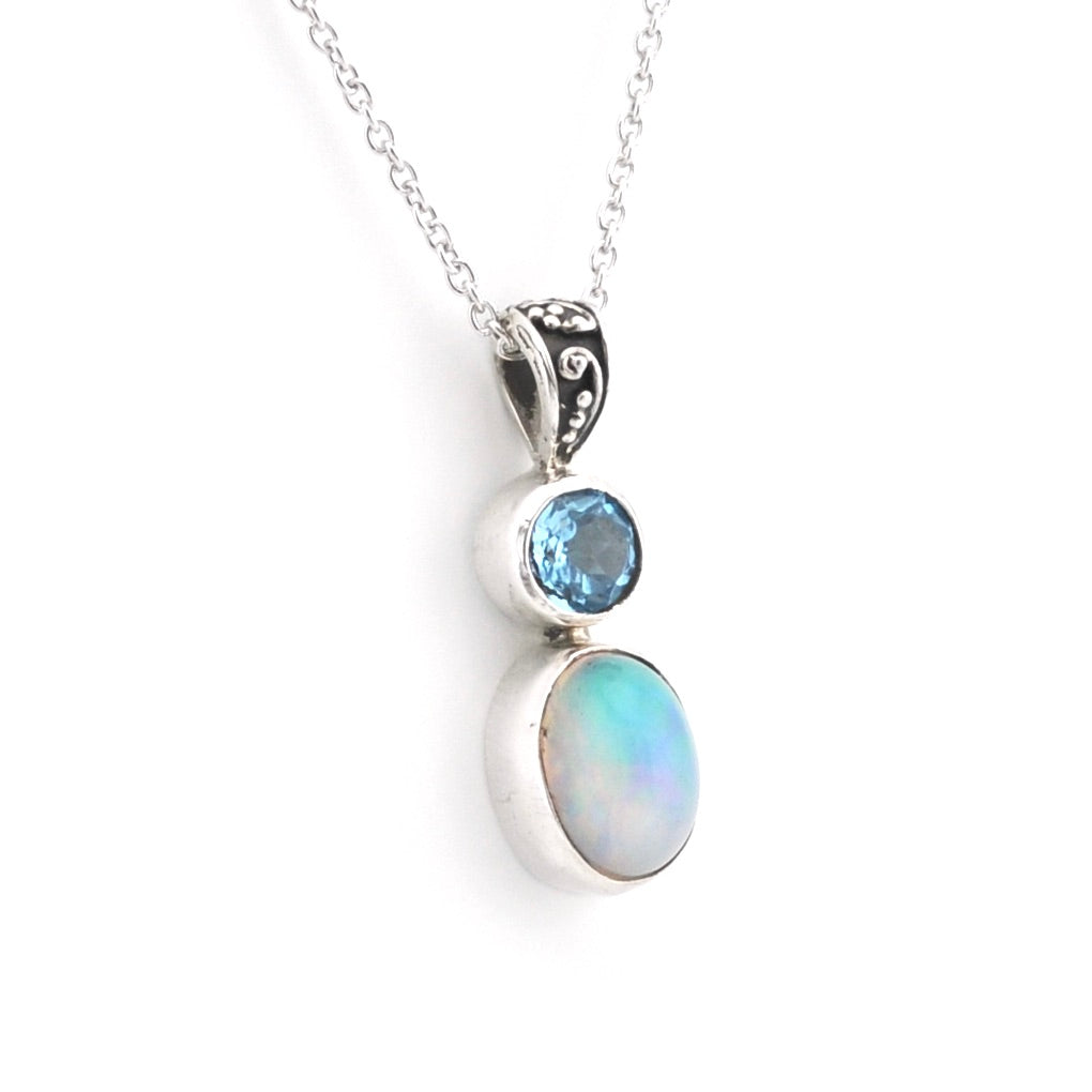 A silver pendant necklace featuring a stacked gemstone design with a small round light blue stone and a larger oval iridescent opal stone, suspended from a delicate silver chain on a white background.