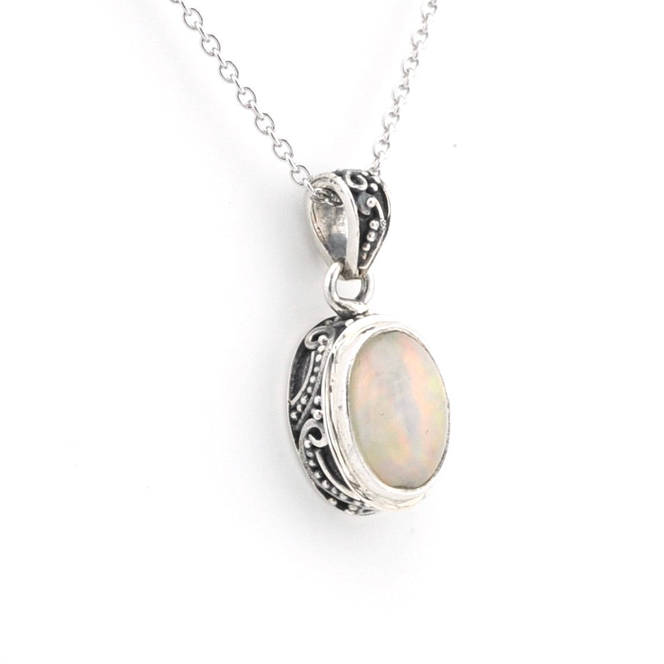 A silver oval pendant necklace featuring a milky white iridescent opal stone, set in an ornate silver bezel with decorative filigree details on the sides, suspended from a delicate silver chain against a plain white background.