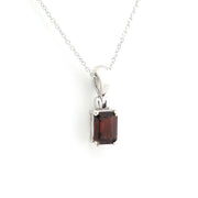 A silver pendant necklace featuring a rectangular emerald-cut dark red garnet gemstone, suspended from a delicate silver chain against a plain white background.