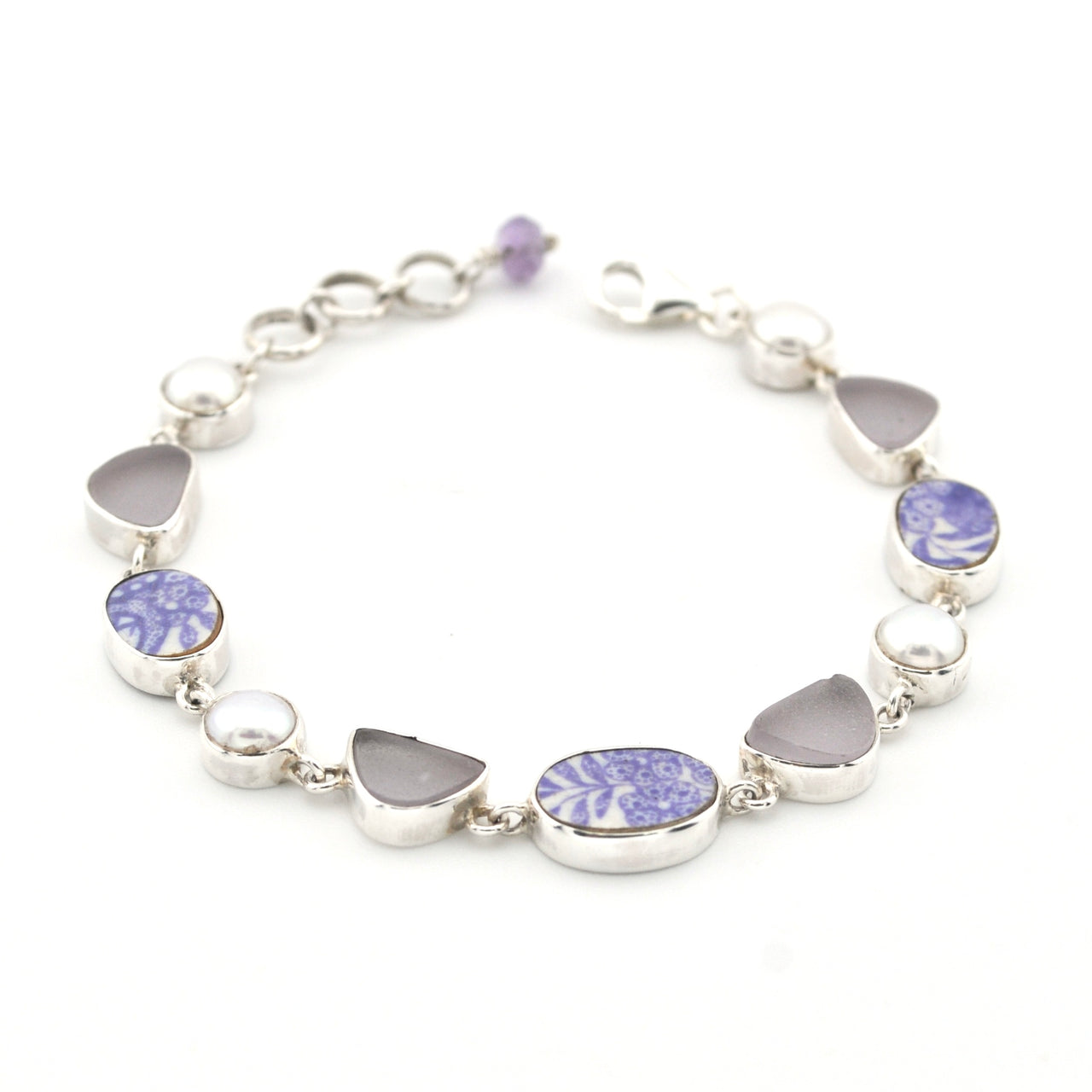 A sterling silver bracelet featuring alternating oval and heart-shaped bezel-set sea glass, including lavender-blue patterned porcelain and pearl on a white background.