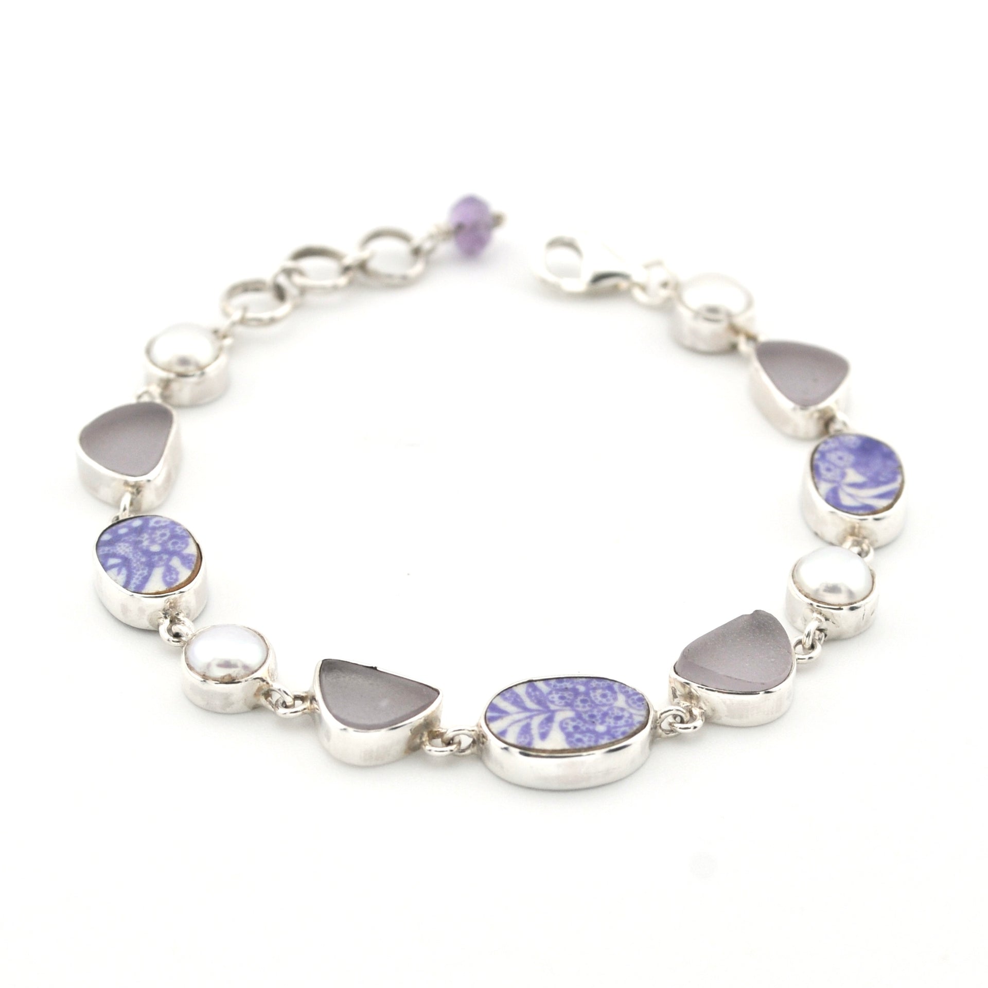 A sterling silver bracelet featuring alternating oval and heart-shaped bezel-set sea glass, including lavender-blue patterned porcelain and pearl on a white background.