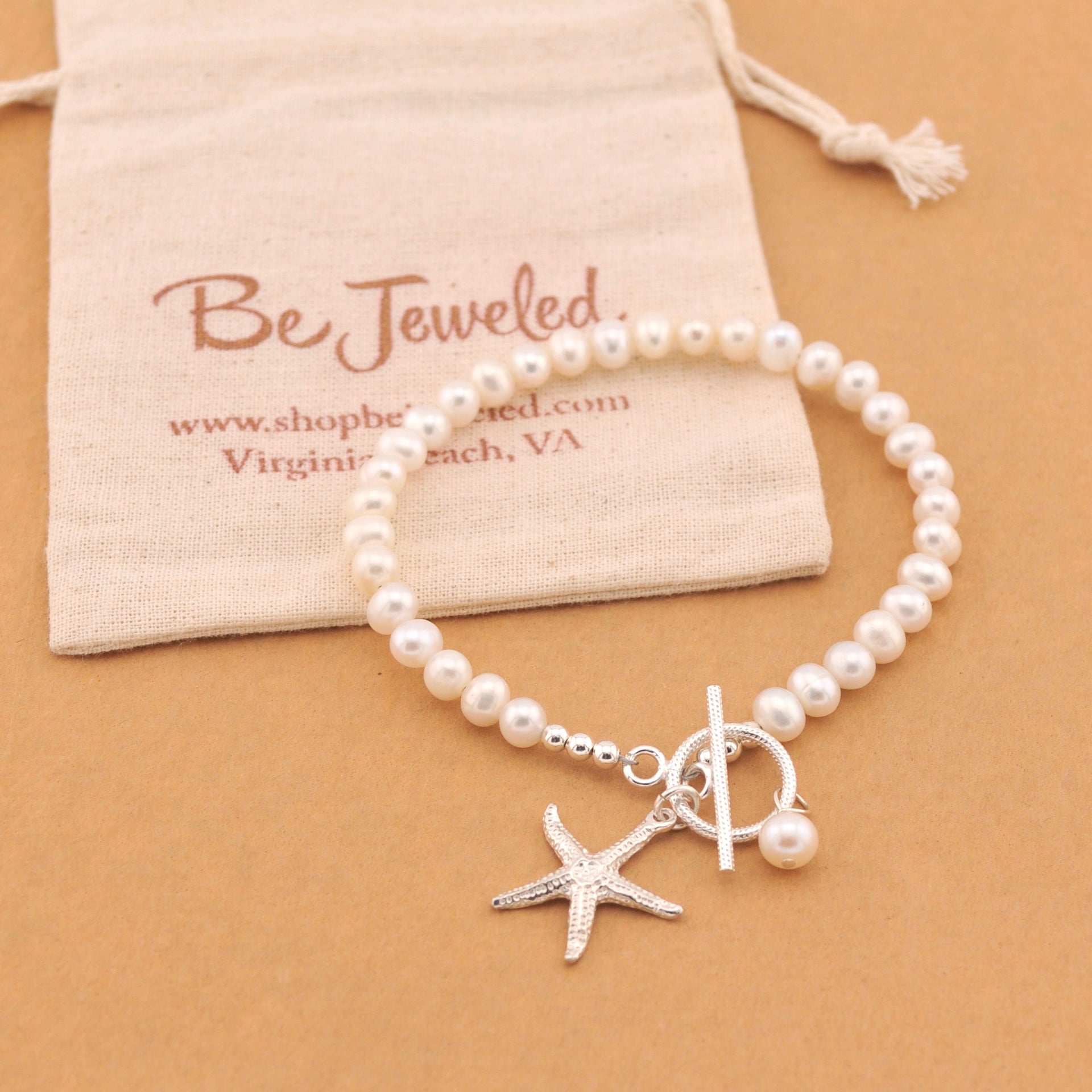 Pearl bracelet with starfish charm on a beige background with 'Be Jeweled' pouch.