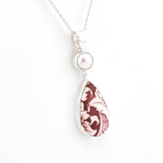 A silver pendant necklace featuring a round white pearl and a teardrop-shaped pendant with a marbled burgundy and white pattern, set against a plain white background.